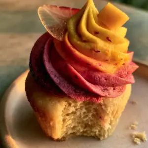 Close-up of a mango strawberry sunset cupcake with a bite missing, revealing a pale gold crumb and tall ombre frosting with fruit flecks.